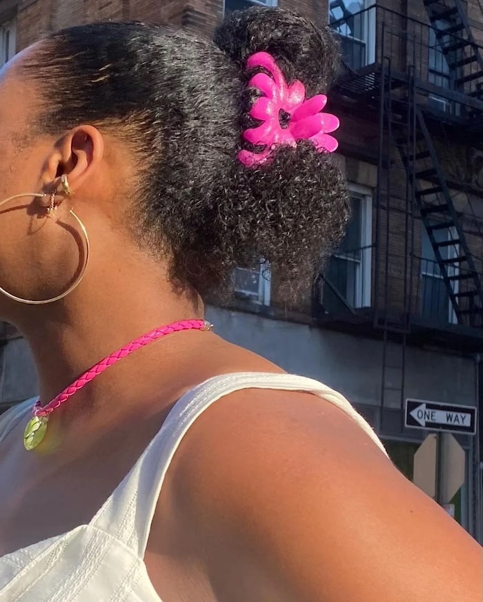 A person with natural curly hair styled in a bun and secured by a bright pink claw clip hair accessory, wearing large hoop earrings, a pink necklace, and a white top, stands outdoors on a summer day near brick buildings and a One Way street sign.