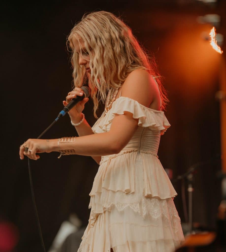 A woman with wavy blonde hair in an off-shoulder, ruffled white dress sings into a microphone on stage under warm lighting, channeling true country star vibes like Ashley Anne.