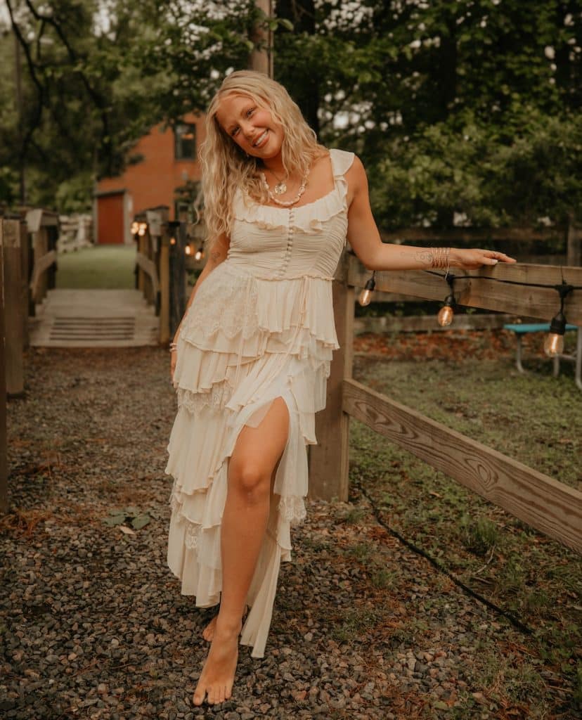 A woman with long blonde hair smiles while standing barefoot on a gravel path. Country star Ashley Anne wears a layered white dress and rests her arm on a wooden fence with string lights, with green trees and a red house in the background.