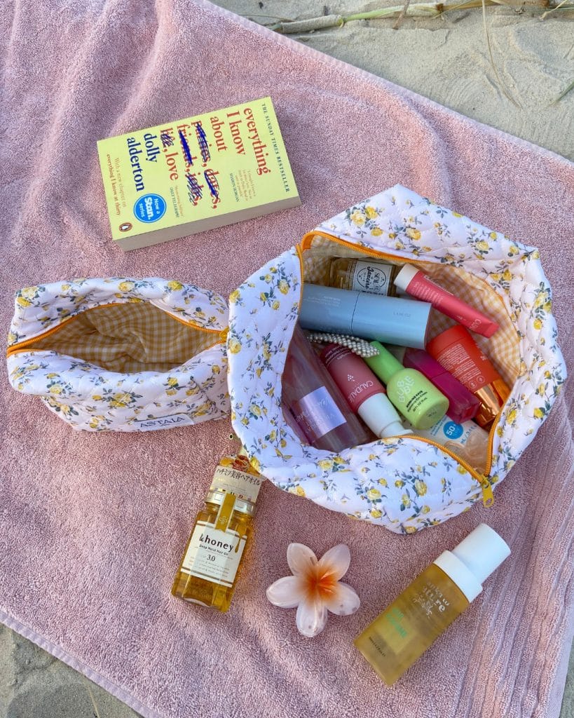 Floral toiletry bags on a pink towel hold various skincare products and cosmetics—perfect beach essentials. Nearby are a book, a flower, face oil, and foaming cleanser, with sand in the background for the ultimate packing guide inspiration.
