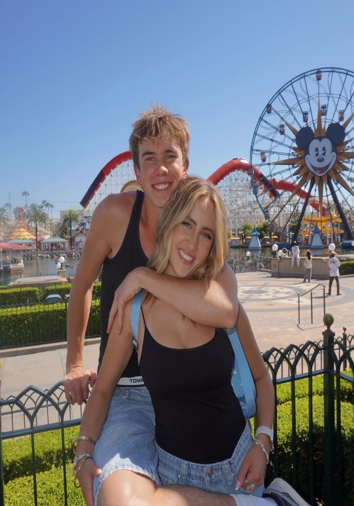 A smiling young couple poses in front of a fence at a theme park, where unexpected places like the Mickey Mouse Ferris wheel and roller coaster make it easy to find a summer crush on a sunny day.