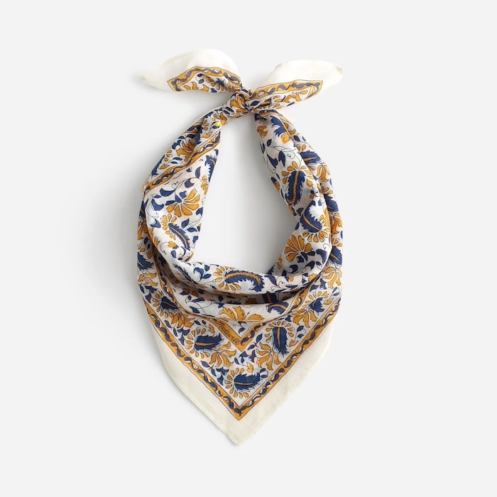 A square scarf with a white background, featuring an intricate floral and leaf pattern in gold and navy blue, tied in a loose knot at the top—perfect as a hair accessory for effortless summer style.
