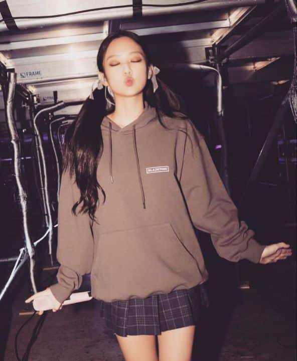 A young woman with long dark hair in pigtails wears a brown hoodie and plaid skirt, standing indoors with eyes closed and lips puckered as if blowing a kiss—like shes prepping backstage for a BLACKPINK DEADLINE world tour. Dim lights, metallic structures behind her.