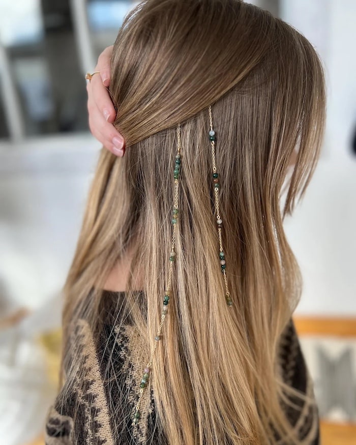 A person with long, straight blonde hair adorned with gold chains and green beads, showcasing a chic summer hair accessory while holding some hair back with one hand. The background is softly blurred indoors.