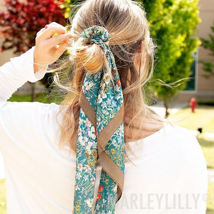 A woman with blonde hair tied in a loose ponytail, accessorized with a long, patterned blue and green floral scarf as a stylish summer hair accessory. She is outdoors in a white top, with trees and a building in the background.