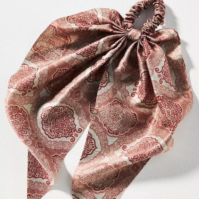 A patterned fabric hair scrunchie with attached scarf tails, featuring a red and beige paisley design, makes a perfect summer hair accessory, laid out on a light background.
