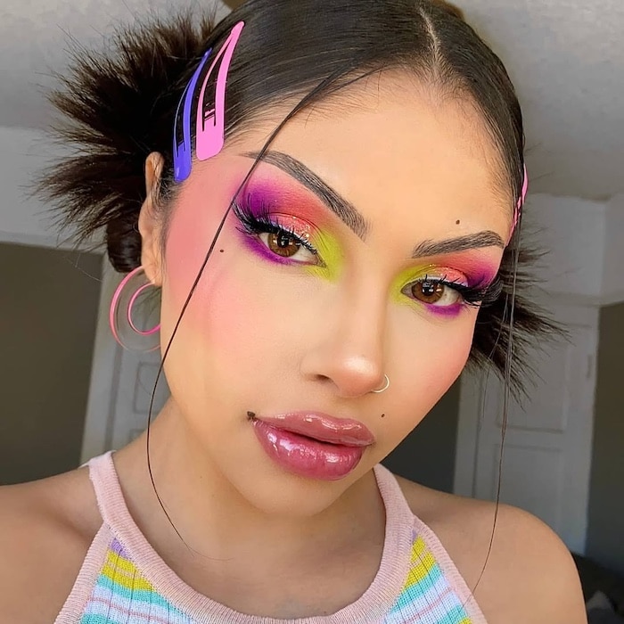 A person with glossy lips, bold colorful eye makeup in pink, orange, and green, and blush. They wear bright pink and purple hair clips, hoop earrings, and a striped pastel top—showcasing vibrant fashion with their hair in two buns.