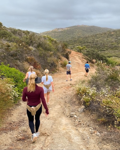 Five people walk along a dirt trail surrounded by green shrubs and hills under a cloudy sky, refreshing their minds and enjoying a relaxing hike in nature.