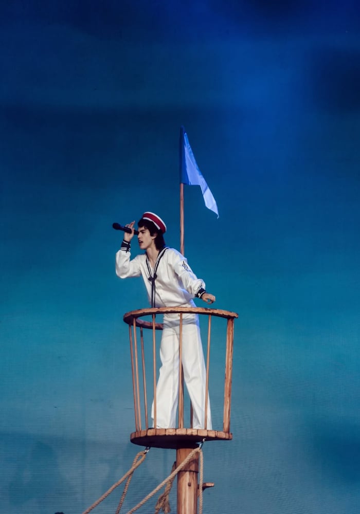 A person dressed in a sailor outfit stands in a ship’s crow’s nest, holding a telescope and looking into the distance, with a blue flag and sky-blue background—capturing celeb moments reminiscent of Gov Ball 2025 adventure.