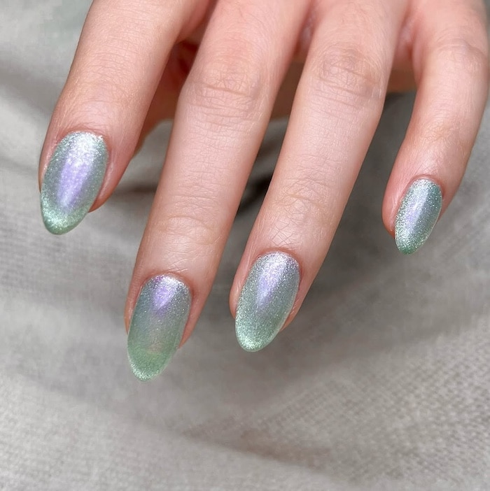 A hand with almond-shaped nails painted in a shimmery, iridescent pastel green and lavender gradient—perfect for a summer manicure. The glossy finish shines against a light, textured fabric background, making this mani trend elegantly eye-catching.