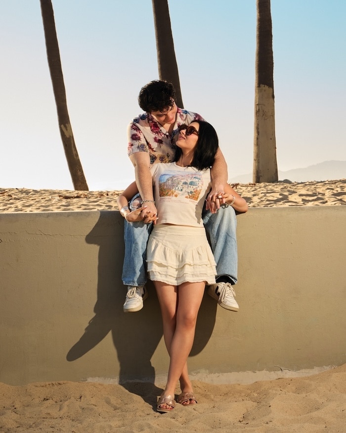 A couple poses affectionately at the beach; Jentzen sits on a low wall, leaning over Elliana, who stands in front of him. Palm trees and sand are visible in the background under a clear blue sky—a true moment for relationship heroes.