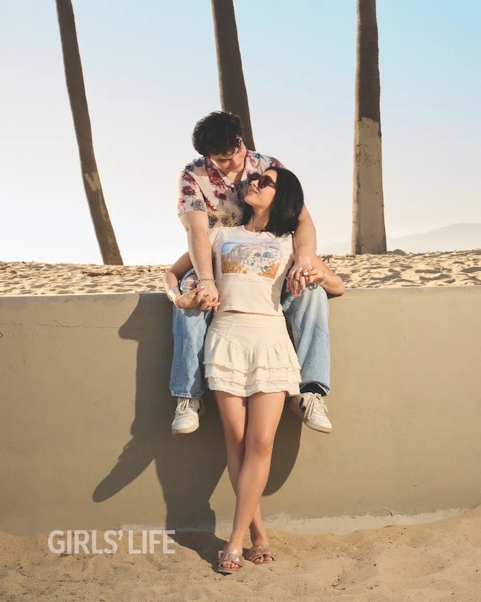 A young couple smiles and poses together at a sunny beach for the Girls Life June/July issue. The man sits on a low wall, leaning over a woman standing in front of him. Both wear casual summer outfits. Palm trees and sand are visible behind them.