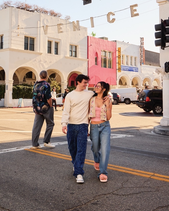 A smiling couple walks arm-in-arm across a street in Venice, California, with the famous Venice sign and colorful buildings in the background. Relationship heroes Jentzen and Elliana lead the way as another person crosses the street behind them.