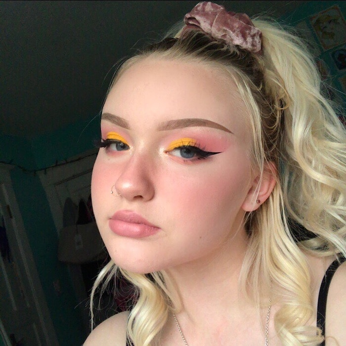 A young person with long blonde hair in a ponytail, wearing a pink scrunchie and vibrant, colorful fashion—pink and yellow eye makeup, winged eyeliner, a nose ring, and small earrings—poses indoors with a neutral expression.