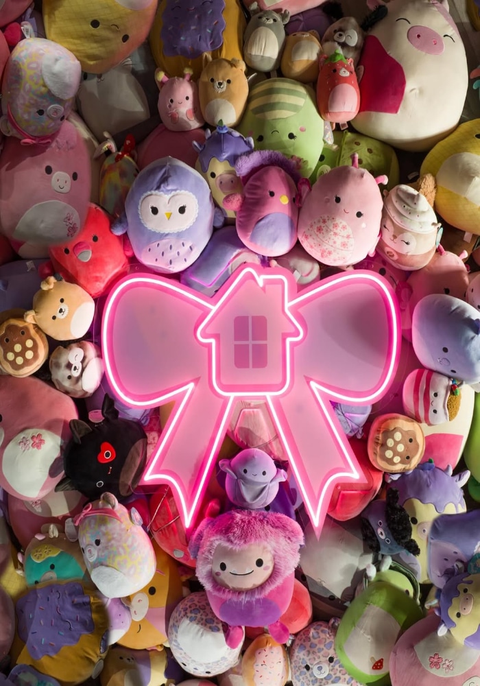 A wall filled with colorful plush toys surrounds a large pink neon sign shaped like a bow with a house icon in the center—an inviting creator house backdrop perfect for TikTok moments. Pastel animals and fantasy creatures add playful charm.