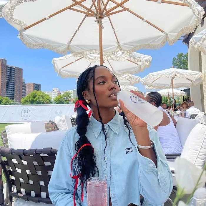 A woman in a blue shirt with red ribbons in her braids, reminiscent of Elliana Walmsley on the Girls Life June/July issue, sits outdoors under white umbrellas, drinking from a clear bottle and holding a pink beverage amid city buildings and greenery.