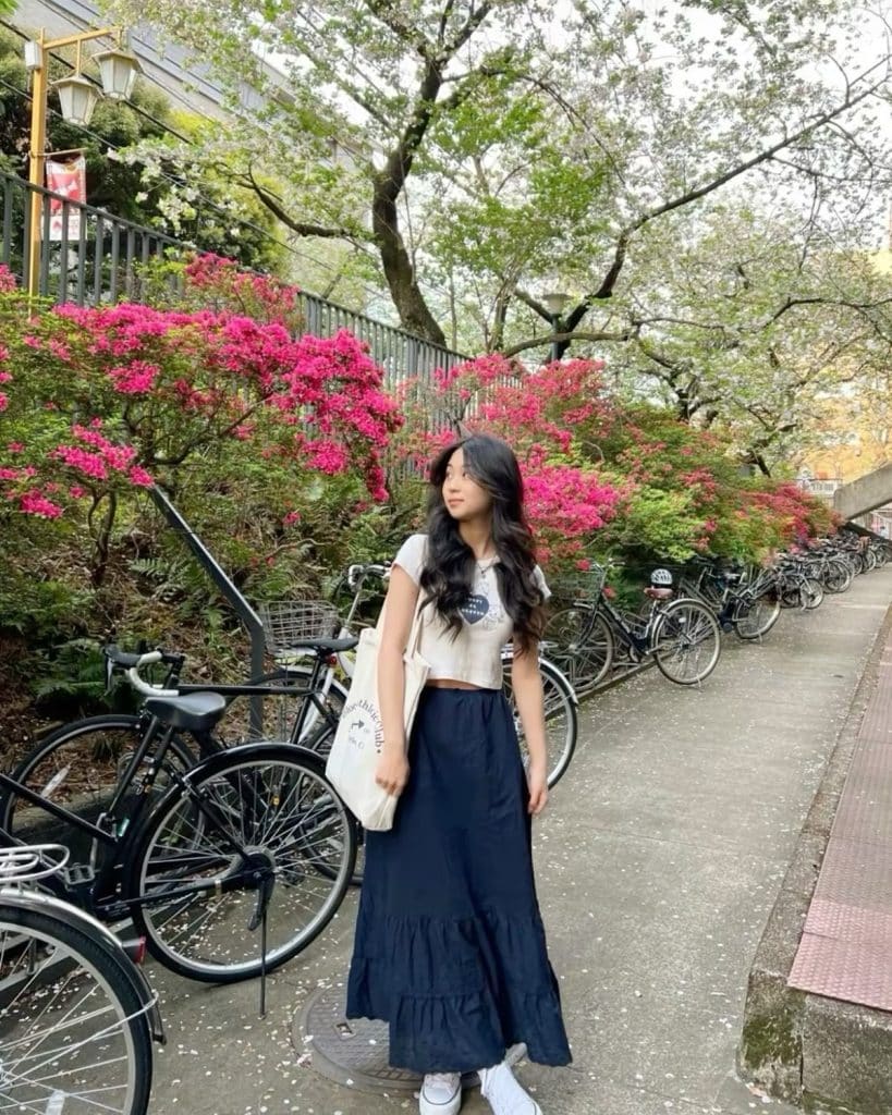 A woman with long dark hair stands on a path lined with parked bicycles, blooming pink flowers, and trees—embracing a summer recharge. She wears a light top, dark skirt, white sneakers, and carries a tote bag, looking off to the side.