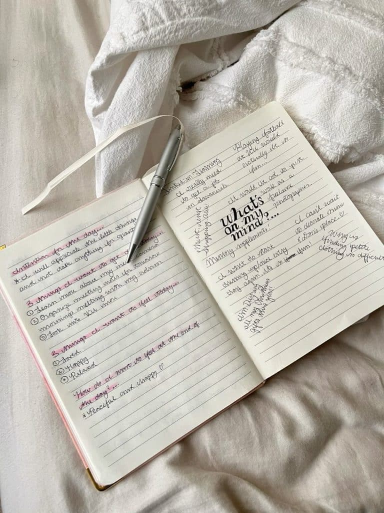 An open journal on a bed with a silver pen resting on it, its pages filled with handwritten notes and prompts like Whats on my mind?—perfect for reflecting on how to recharge during your summer recharge. A white blanket is partially visible.