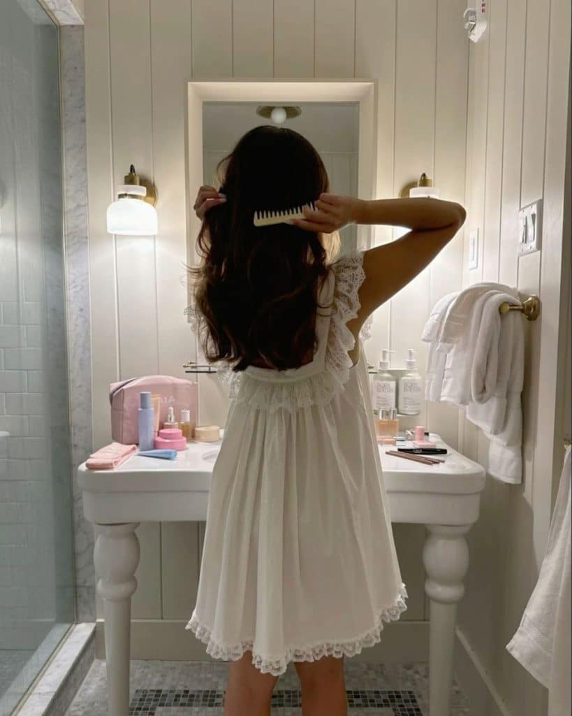 A woman in a white nightdress stands in a bathroom, facing away, brushing her long hair in front of a sink with various skincare and beauty products arranged on the counter for a summer recharge.