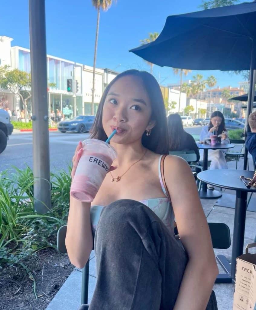 A young woman enjoys a summer recharge at an outdoor café, sipping a pink drink through a straw. Casually dressed with one leg up, she relaxes among greenery, bustling storefronts, and other people on a sunny day.