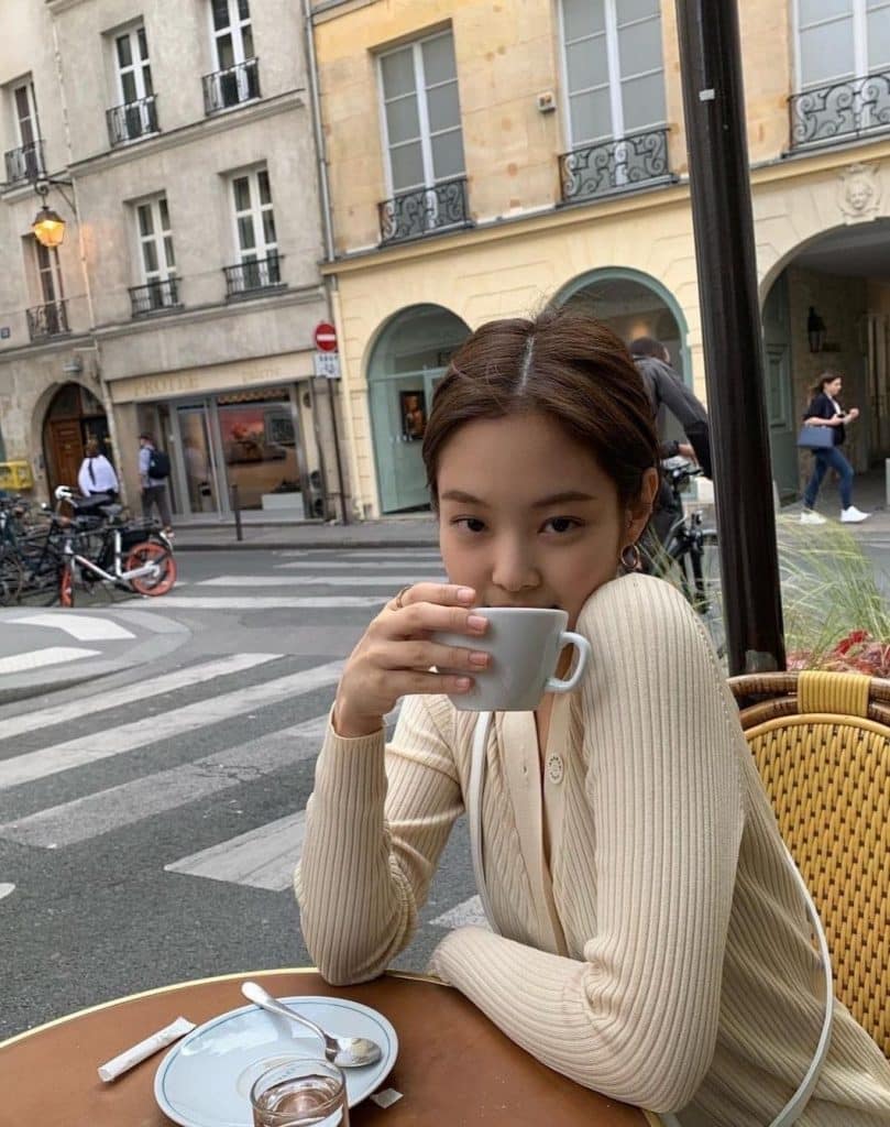 A young woman enjoys a summer recharge at an outdoor café, holding a white cup close to her face and looking at the camera. She is wearing a light-colored ribbed sweater with city streets, bikes, and buildings in the background.