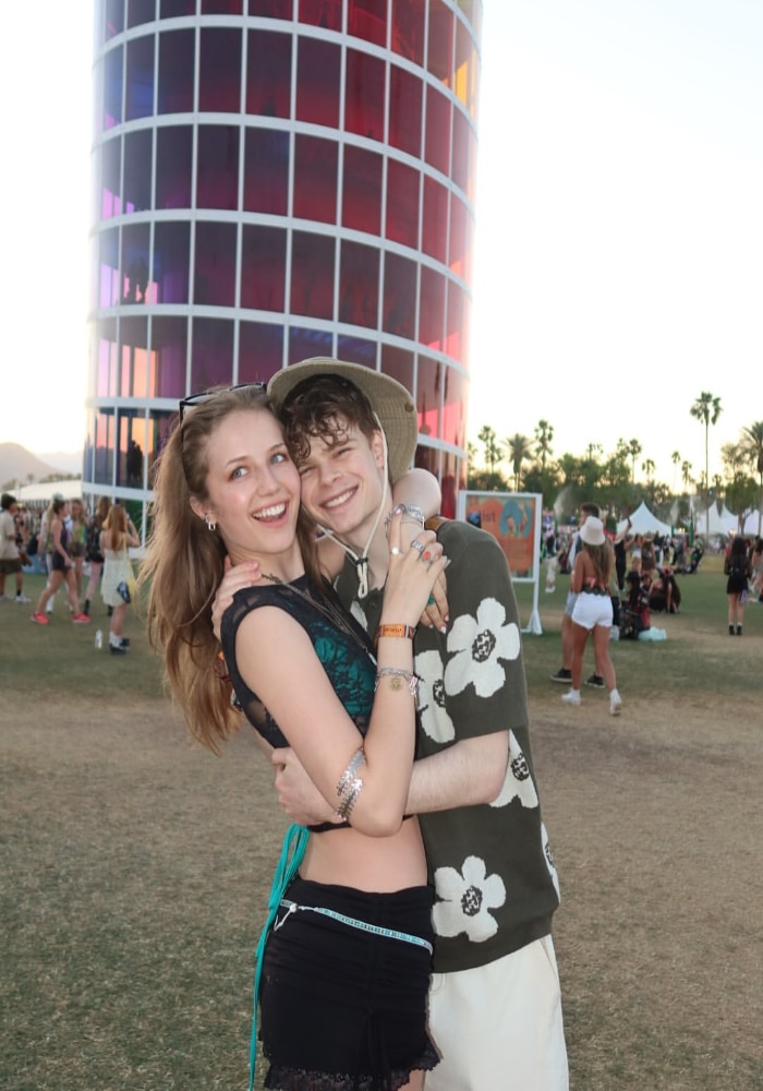 A young couple smiles and embraces in front of a colorful, cylindrical tower at an outdoor festival—proof you can find a summer crush in the most unexpected places. Palm trees and cheerful crowds complete the sunny scene.