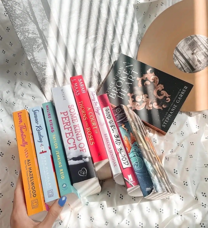 A hand holds a row of colorful books arranged on a bed with a white patterned sheet. Sunlight streams in, casting shadows. A vinyl record and its sleeve are nearby, along with the June/July issue of Girls Life featuring Elliana Walmsley.