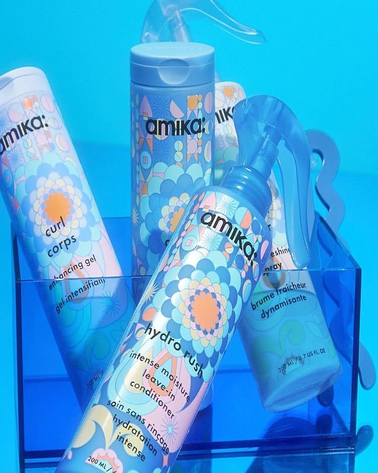 Four colorful Amika hair care products, including a spray bottle and tubes, are displayed in a clear blue acrylic container against a bright blue background. The packaging features pastel geometric floral designs—perfect for hydrated curls or curly hair care.