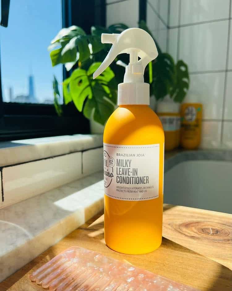 A yellow bottle of milky leave-in conditioner for hydrated curls sits on a bathroom counter next to a pink comb, with sunlight streaming through a window and a green potted plant in the background.