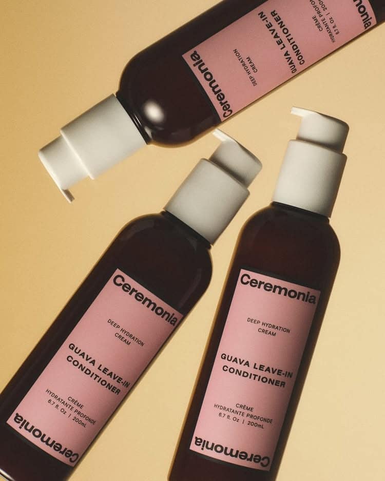Three brown bottles of Ceremonia Guava Leave-In Conditioner with pink labels are arranged diagonally on a beige background. Each bottle has a white pump dispenser and black text, perfect for keeping curls hydrated and defined.