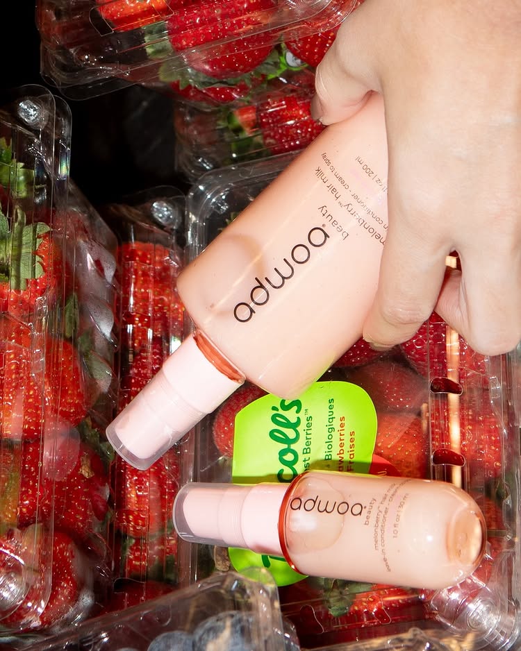 A hand holds a pink Adwoa Beauty leave-in conditioner bottle above clear containers of fresh strawberries, with another Adwoa bottle and a green Driscoll’s strawberry label visible—perfect for hydrated curls.