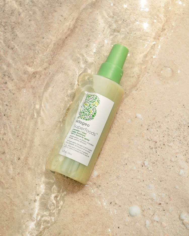 A green and white bottle of Briogeo Superfoods hair product lies on a sandy beach, partially covered by a gentle stream of water—perfect for maintaining hydrated curls even in sun and surf.