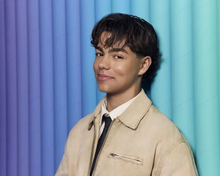Elliana Walmsley, with short, wavy brown hair, wears a beige jacket and black tie, smiling slightly while standing in front of a gradient blue and purple backdrop for the June/July issue of Girls Life.