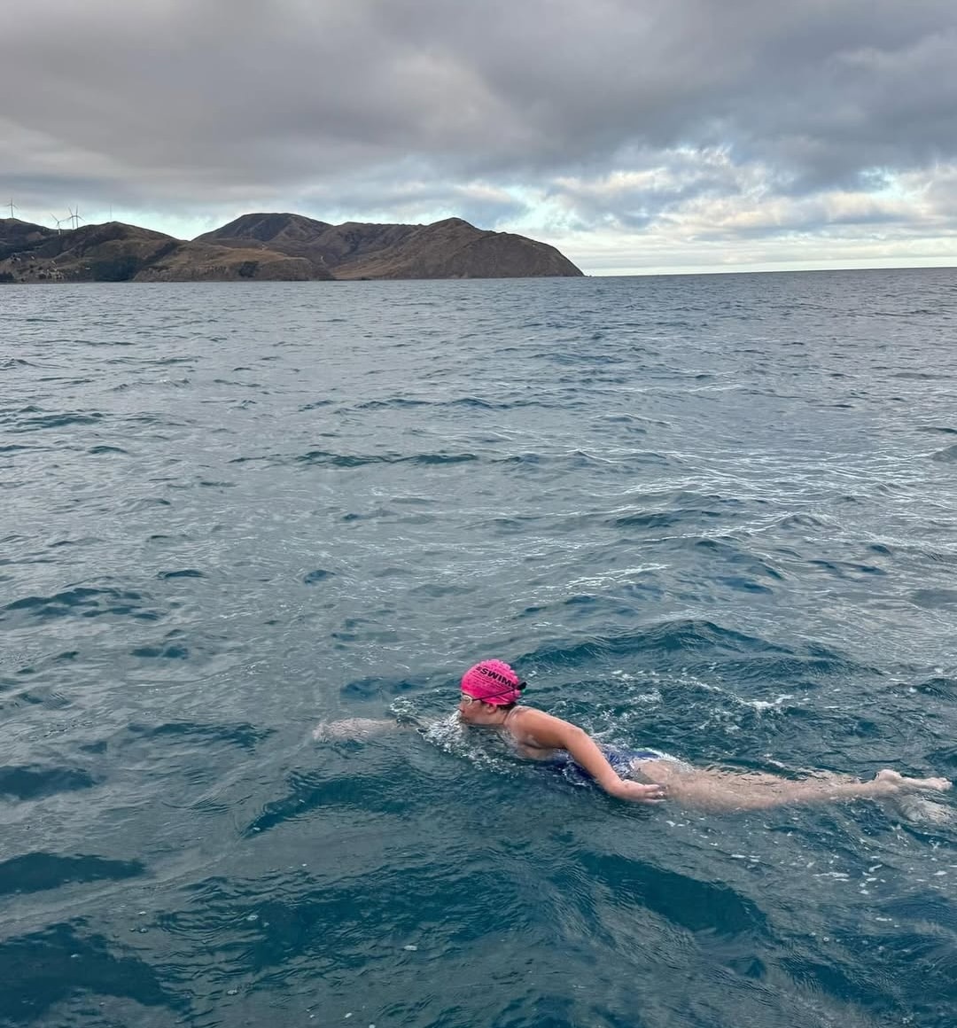 A 17-year-old rockstar wearing a pink swim cap and swimsuit swims freestyle in open water near a rugged coastline, under a cloudy sky with distant hills—channeling the spirit of those who have swam the Cook Strait.