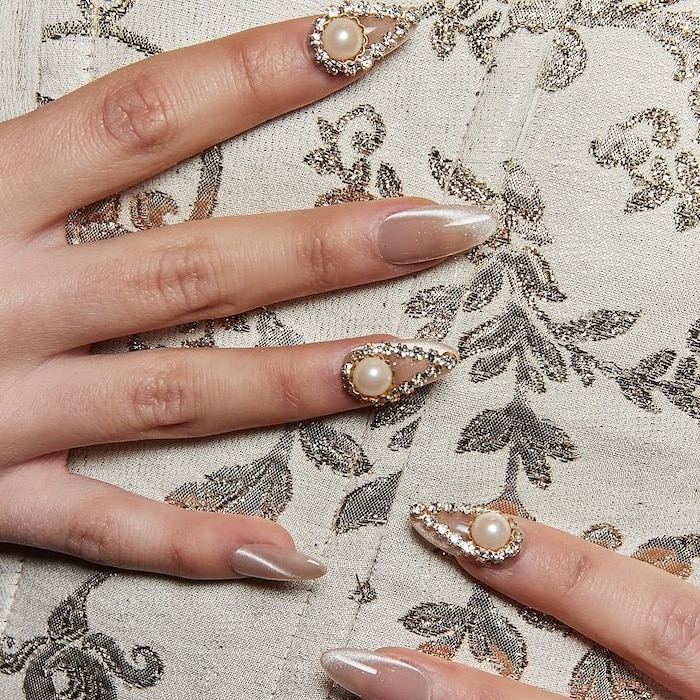 Close-up of a hand with long, almond-shaped nails showcasing a summer manicure. Some nails are decorated with pearls and rhinestones, while others feature a glossy nude color. The hand rests on cream patterned fabric, highlighting this chic mani trend.