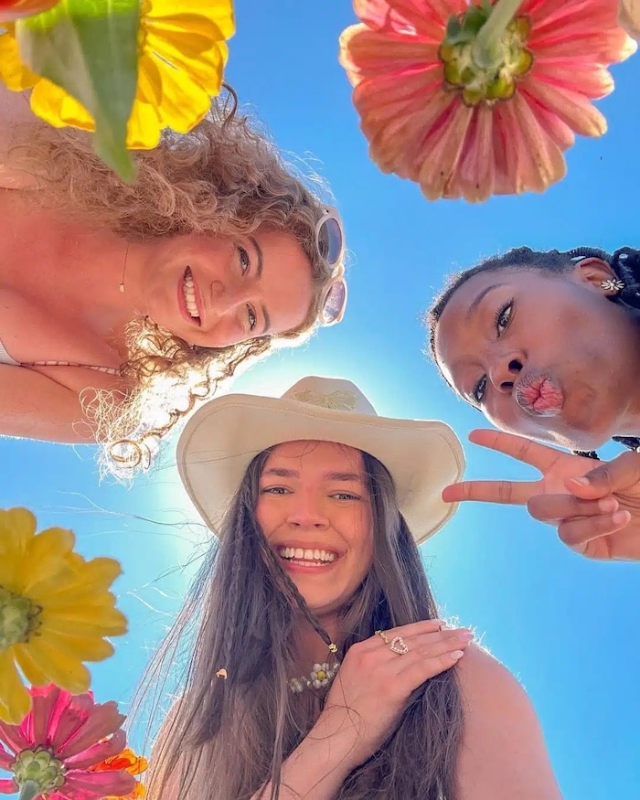 Three women smiling and posing from a low angle, surrounded by colorful flowers and a bright blue sky. One makes a peace sign and wears a white hat, capturing the fun vibe of a Girls Life June/July issue cover.