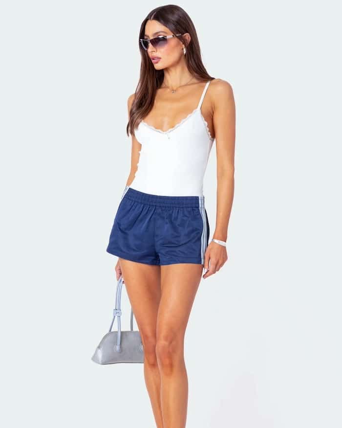 A woman wearing a white camisole, navy blue athletic shorts, sunglasses, and carrying a small silver handbag stands against a plain light background—a chic look inspired by 4th of July fashion style guide trends.