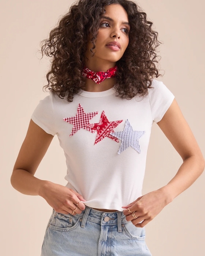 A woman with curly hair showcases 4th of July fashion in a white t-shirt with three checkered star patches, light blue jeans, and a red bandana around her neck. She poses confidently with one hand in her pocket against a neutral background.