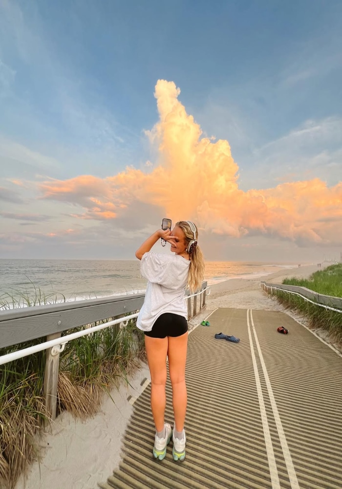 A woman in a white t-shirt and black shorts stands on a beach boardwalk, capturing dramatic orange clouds at sunset—an inspiring scene for anyone on a summer shape-up or fun workout journey. Sand, grass, and scattered belongings are visible nearby.