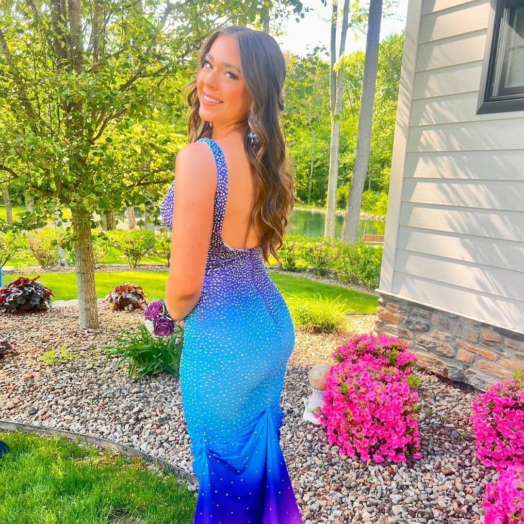A young woman in a sparkly blue and purple gradient gown poses and smiles outdoors near a garden with pink flowers, trees, and a pond, next to a white house—perfect inspiration for prom dress shopping.