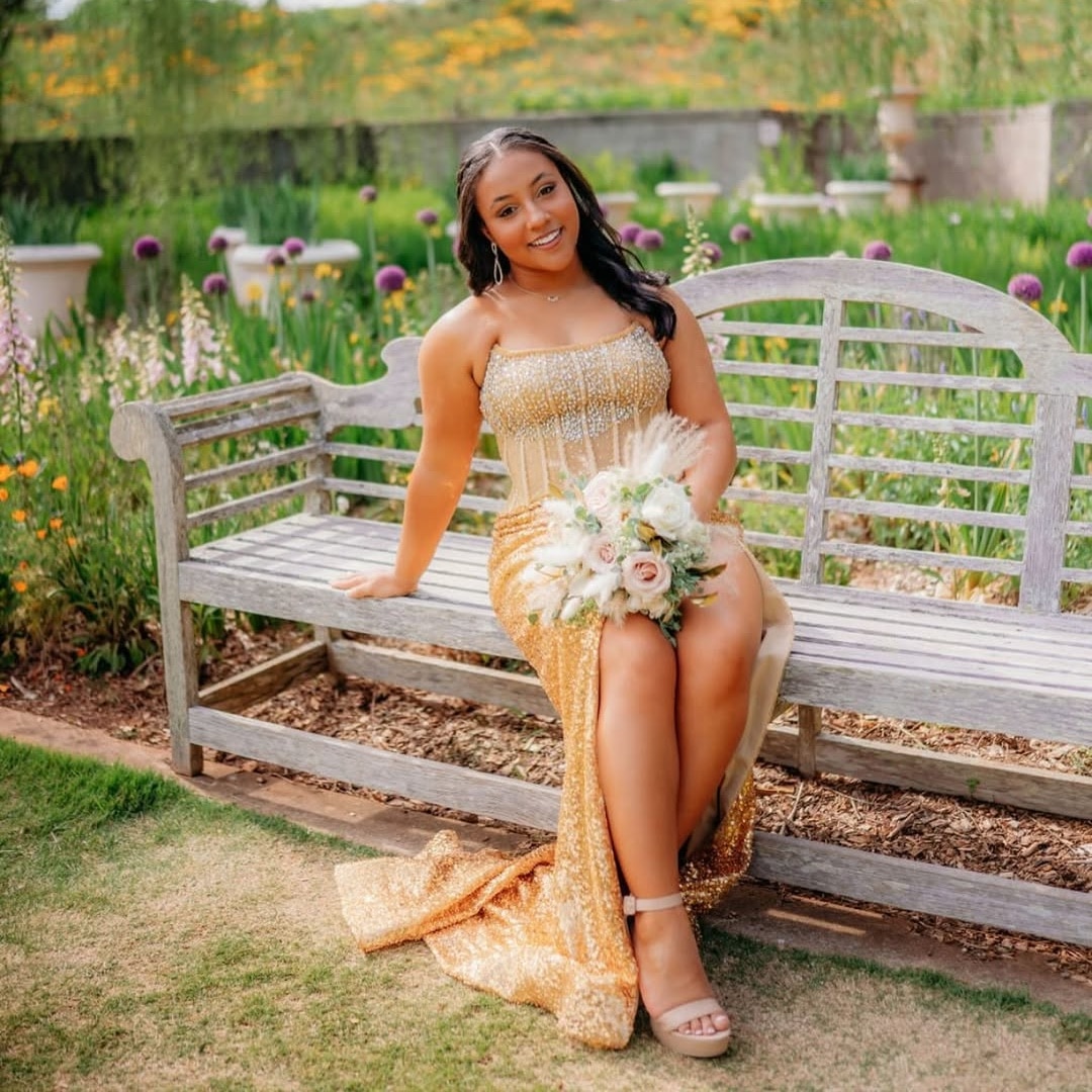 A young woman in a sparkling gold prom dress sits on a wooden bench in a garden, smiling and holding a bouquet of flowers. Colorful blooms and greenery fill the background, capturing the joy of prom dress shopping.