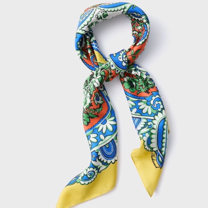 A silk scarf with a colorful, intricate pattern featuring blue, green, yellow, red, and white designs, tied in a loose knot as a chic summer hair accessory against a plain light background.