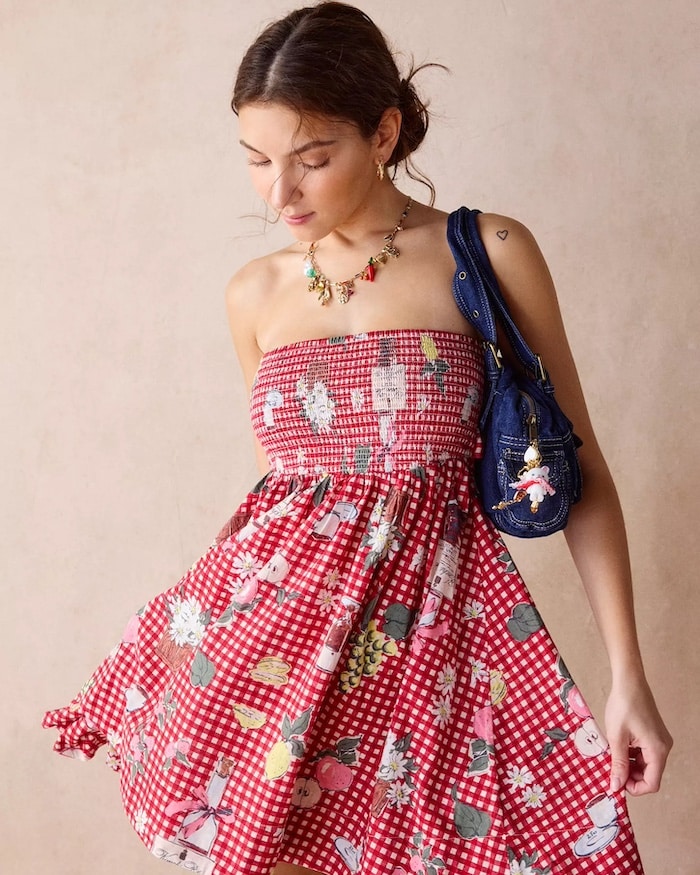 A woman wearing a red gingham strapless dress with a floral print—perfect for a 4th of July style guide—looks down, holding one side of the skirt. She has a blue shoulder bag and a colorful necklace, with her brown hair tied back.