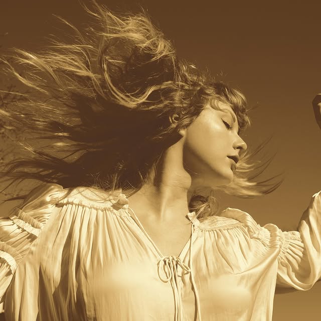 A woman in a flowing, light-colored blouse stands outdoors with her eyes closed and hair blowing in the wind, bathed in warm, sepia-toned light—evoking the nostalgic mood of Taylor Swift’s Vault Tracks.