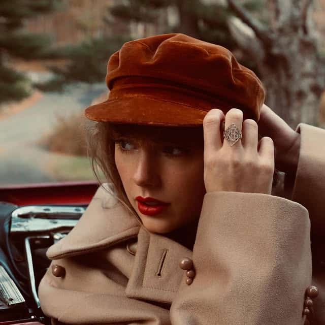 A woman in a beige coat and brown cap sits in a car, adjusting her hat. She has wavy hair, red lipstick, and wears a large ring—her style reminiscent of Taylor Swift songs. Trees and a road blur softly in the background.