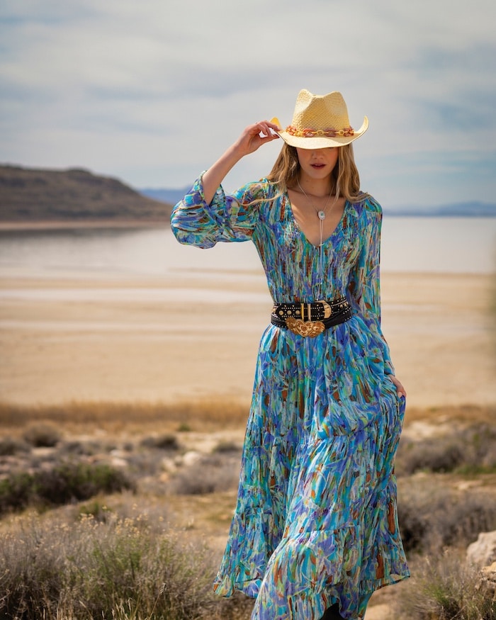 A woman in a flowing, colorful blue maxi dress and straw hat stands outdoors by a sandy beach with hills in the background, enjoying the warmth of the sun and the promise of solar energy as she holds her hat and looks down.
