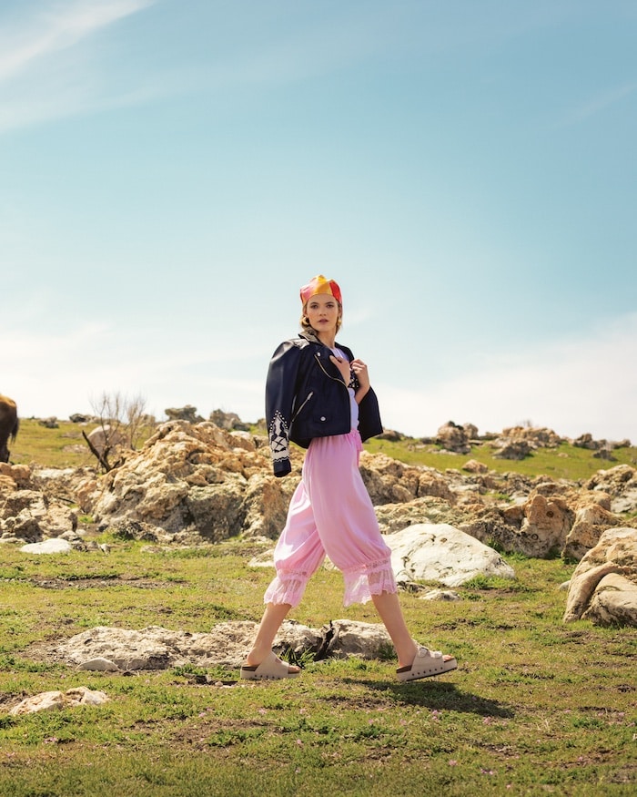 A woman wearing a pink dress, dark jacket, and headscarf walks on grassy, solar powered terrain dotted with rocks beneath a blue sky.