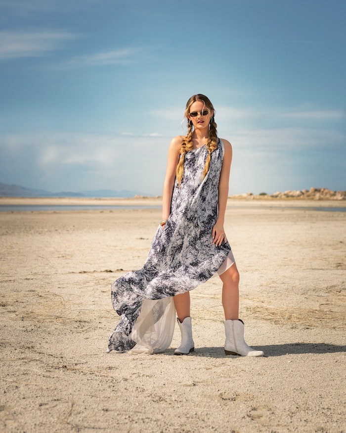 A woman with long braided hair wearing sunglasses, a flowing black and white patterned dress, and white boots stands on a dry, sandy landscape under the bright blue sky, embracing the power of solar energy.