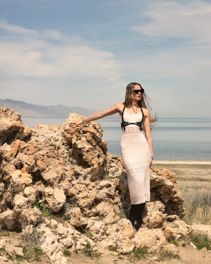 A woman in a white patterned dress and sunglasses stands by large rocky formations near a body of water, with solar-powered lanterns lighting the scene and mountains in the background under a partly cloudy sky.