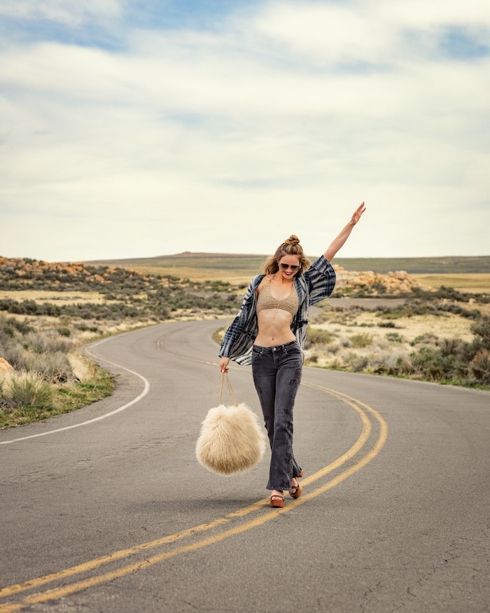 A woman strolls down a winding road in a rural landscape, smiling with one arm raised. Sporting a crop top, open shirt, dark pants, sandals, and sunglasses, she carries a large solar powered fluffy bag under the partly cloudy sky.